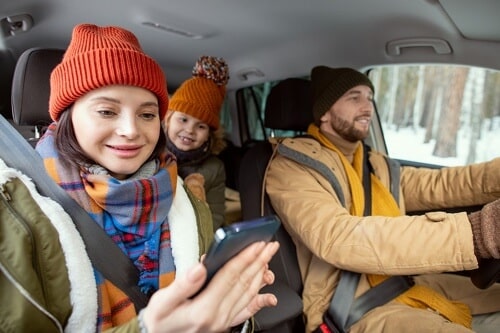 Safe Holiday Driving Tips For Drivers in Centerville, OH with C's Autohaus image of dad, mom, and daughter in winter clothes driving in the car while mom looks at her smart phone for directions