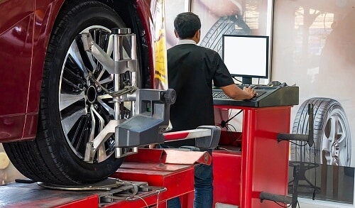 Wheel Alignment by C’s Autohaus in Centerville, OH. Closeup image of a car on stand with sensors on wheels for wheels alignment, with mechanic operating the alignment machine on the background.