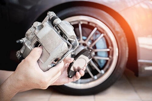 Brake Repair & Brake Inspection in Centerville, OH | C’s Autohaus. Image of a man’s hand holding brake calliper and brake pad spare parts. At the background is a car tire.