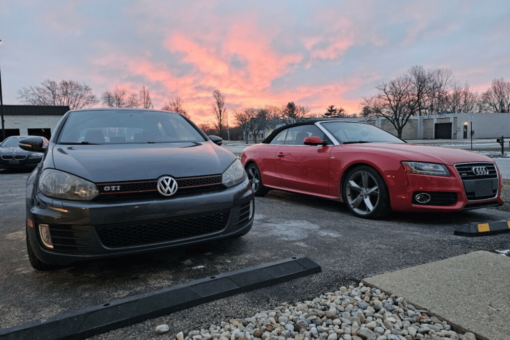 european car repair, auto repair in Centerville, Ohio. C's Autohaus. A black Volkswagen GTI and a red Audi convertible parked side by side at sunset, with a vibrant sky in the background.