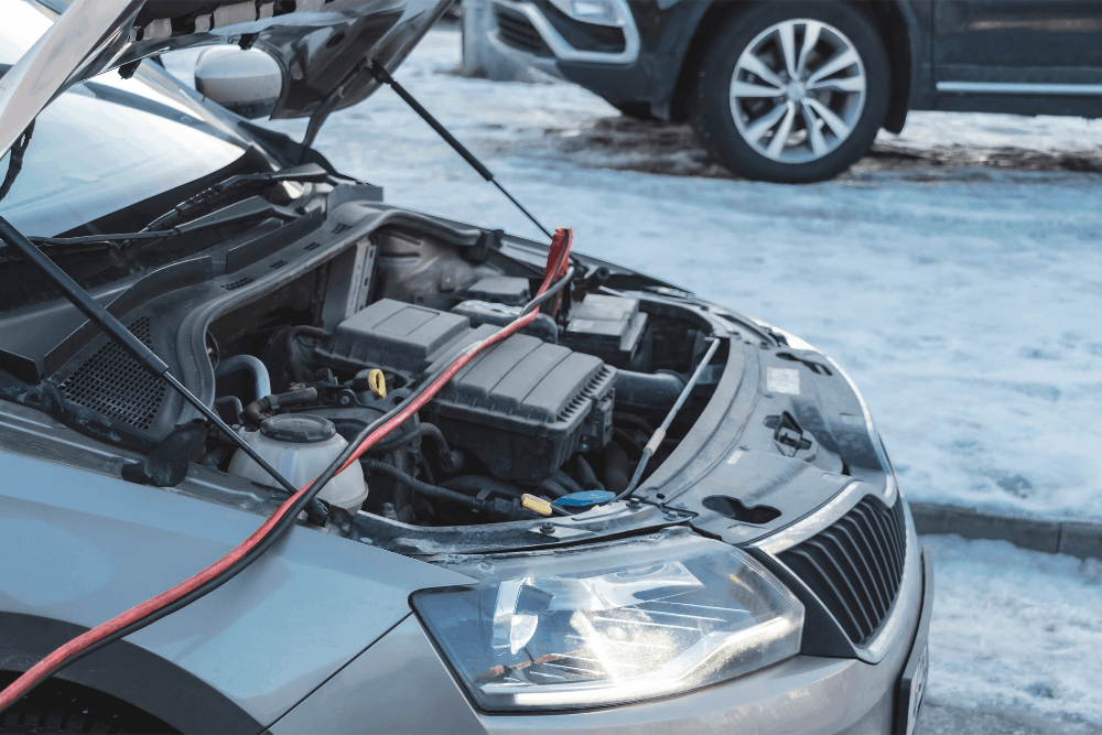 Car battery failure, auto repair in Centerville, OH by C’s Autohaus. Image of a sedan with the hood open and jumper cables connected during a cold winter day, emphasizing reliable battery assistance and emergency starting support.